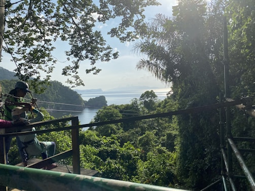 A lush tropical landscape with dense greenery spreads out beneath a clear sky. In the foreground, a person is preparing to enjoy a zipline ride, secured in a harness and helmet. The zipline cable extends over the verdant treetops toward water in the distance. Sunlight filters through the foliage, creating a serene and adventurous atmosphere.