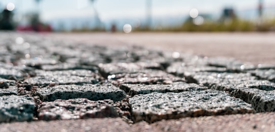 A detailed and close-up view of a cobblestone pavement under natural light. The image is focused on the texture and arrangement of the cobblestones, with a shallow depth of field blurring the background. In the blurred background, there are hints of greenery and the sky.