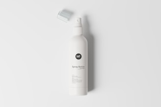 A minimalistic spray bottle lies horizontally on a plain white background. The bottle is cylindrical with a sleek design and a removable cap placed nearby. The label has a logo and the text 'Spray Bottle'.