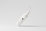 A white spray bottle with a minimalistic design is depicted against a light gray background. The bottle's label includes stylized text and a logo, and its cap is suspended in mid-air, suggesting motion.