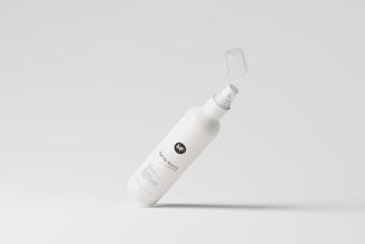 A white spray bottle with a minimalistic design is depicted against a light gray background. The bottle's label includes stylized text and a logo, and its cap is suspended in mid-air, suggesting motion.