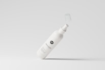 A white spray bottle with a minimalistic design is depicted against a light gray background. The bottle's label includes stylized text and a logo, and its cap is suspended in mid-air, suggesting motion.