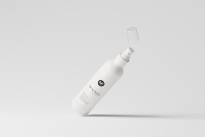 A white spray bottle with a minimalistic design is depicted against a light gray background. The bottle's label includes stylized text and a logo, and its cap is suspended in mid-air, suggesting motion.