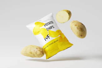 A fresh green bag of tomato banana chips floating gently above a soft gray surface, surrounded by floating chips and slices.