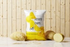A package of potato chips is placed on a marble surface in front of a wooden panel background. Two whole potatoes and one sliced potato are positioned next to the bag. The package design features realistic images of potato chips and text branding.