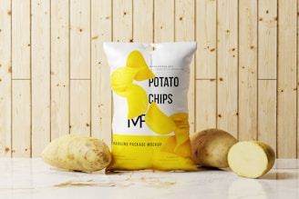 A package of potato chips is placed on a marble surface in front of a wooden panel background. Two whole potatoes and one sliced potato are positioned next to the bag. The package design features realistic images of potato chips and text branding.