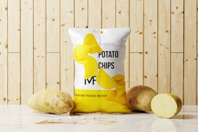 A package of potato chips is placed on a marble surface in front of a wooden panel background. Two whole potatoes and one sliced potato are positioned next to the bag. The package design features realistic images of potato chips and text branding.
