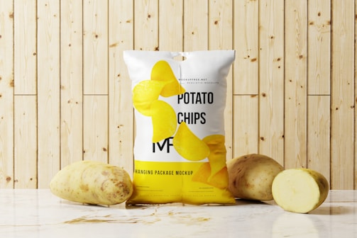 A package of potato chips is placed on a marble surface in front of a wooden panel background. Two whole potatoes and one sliced potato are positioned next to the bag. The package design features realistic images of potato chips and text branding.