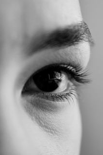 Close-up of a human eye in black and white, focusing on the eyelashes and eyebrow with detailed texture of the skin visible.