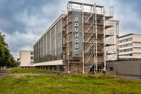 A large, modernist building with a distinctively clean, geometric style set against a cloudy sky. The front of the building features a prominent vertical sign labeled 'Bauhaus,' and the structure is surrounded by scaffolding, suggesting ongoing renovation. The architecture includes expansive glass windows and white walls. A grassy area with a few wildflowers is visible in the foreground, and there are other similar buildings in the background along a tree-lined street.