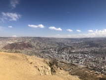 A panoramic view of a city nestled in a mountainous terrain under a vast, clear blue sky scattered with a few fluffy clouds. The city is densely populated with numerous buildings, and the surrounding hills have a rugged texture with patches of greenery and earth-toned rock formations.