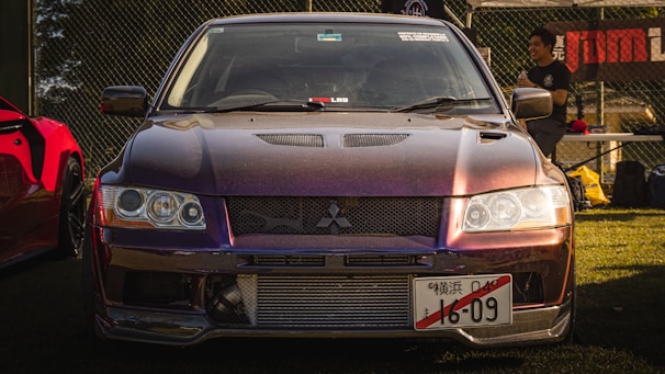 A modified Mitsubishi sports car is prominently displayed, featuring a sleek, angular front design and a reflective, dark-colored finish. The vehicle is parked on grass near a chain-link fence with a banner in the background. A person stands near another car with a red color on the left, engaged in conversation.