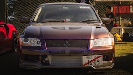 A modified Mitsubishi sports car is prominently displayed, featuring a sleek, angular front design and a reflective, dark-colored finish. The vehicle is parked on grass near a chain-link fence with a banner in the background. A person stands near another car with a red color on the left, engaged in conversation.