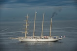 A large, traditional sailing ship with three masts is on calm waters under a cloudy sky. Smoke is billowing from the rear, indicating the use of an onboard engine or generator. The ship features a white hull and appears to be equipped for long-distance travel.