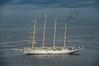 A large, traditional sailing ship with three masts is on calm waters under a cloudy sky. Smoke is billowing from the rear, indicating the use of an onboard engine or generator. The ship features a white hull and appears to be equipped for long-distance travel.