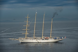 A large, traditional sailing ship with three masts is on calm waters under a cloudy sky. Smoke is billowing from the rear, indicating the use of an onboard engine or generator. The ship features a white hull and appears to be equipped for long-distance travel.
