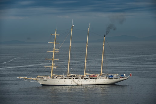 A large, traditional sailing ship with three masts is on calm waters under a cloudy sky. Smoke is billowing from the rear, indicating the use of an onboard engine or generator. The ship features a white hull and appears to be equipped for long-distance travel.