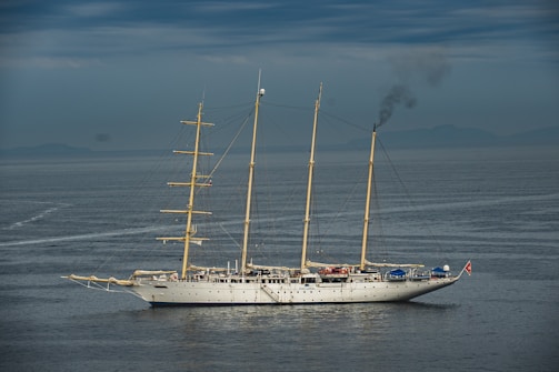 A large, traditional sailing ship with three masts is on calm waters under a cloudy sky. Smoke is billowing from the rear, indicating the use of an onboard engine or generator. The ship features a white hull and appears to be equipped for long-distance travel.