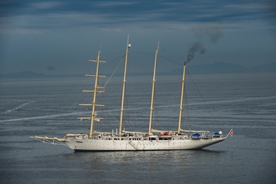 A large, traditional sailing ship with three masts is on calm waters under a cloudy sky. Smoke is billowing from the rear, indicating the use of an onboard engine or generator. The ship features a white hull and appears to be equipped for long-distance travel.
