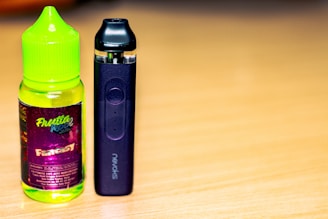 A bottle of fruit-flavored vape juice with a bright green cap and colorful label sits next to a sleek black vape device on a wooden surface.