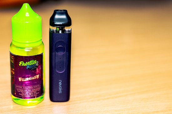 A bottle of fruit-flavored vape juice with a bright green cap and colorful label sits next to a sleek black vape device on a wooden surface.
