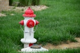 A red and white fire hydrant is situated on a lush green lawn. The hydrant is accented with metal chains and has a small patch of bare soil beside it. In the background, there are some stone gravel and a hint of a brick structure.