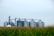 Several large grain silos are visible in the background with interconnected metal structures above them. In the foreground, there is a field of tall green corn.