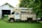 An old utility truck is parked on a grassy area beside a white wooden garage with overgrown greenery. The vegetation partially obscures the truck's tires and parts of the garage. A green tractor is visible inside the garage, while the foliage in the background suggests a rural setting.
