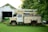 An old utility truck is parked on a grassy area beside a white wooden garage with overgrown greenery. The vegetation partially obscures the truck's tires and parts of the garage. A green tractor is visible inside the garage, while the foliage in the background suggests a rural setting.
