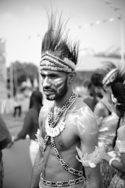 A man stands in traditional attire, adorned with beads and wearing tribal makeup and a headdress. His expression is serious, and his hair is styled upward in a striking fashion. Other people in similar attire are blurred in the background, suggesting a cultural or festive gathering.