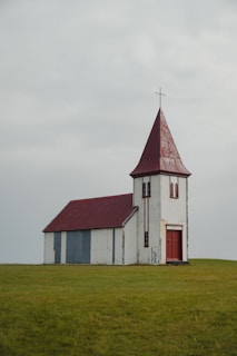 A small, rustic church with a red roof and white wooden walls stands alone in a vast grassy field. The church has a tall spire topped with a metal cross. The structure appears weathered, adding a sense of age and simplicity to the scene.
