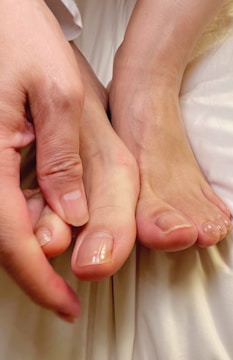 A close-up view of feet resting on a soft, light-colored fabric, with a hand gently touching the side of the foot. The toenails appear clean and well-maintained.