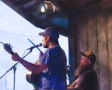Two men are performing music on a wooden stage, one playing a guitar and singing into a microphone, while the other is playing a different instrument and wearing a cap and has a thick beard. The scene appears to be happening under an overhang or roofed area, possibly during dusk or early evening.