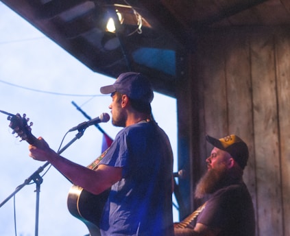 Two men are performing music on a wooden stage, one playing a guitar and singing into a microphone, while the other is playing a different instrument and wearing a cap and has a thick beard. The scene appears to be happening under an overhang or roofed area, possibly during dusk or early evening.