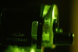 A close-up view of a mechanical or optical device featuring a metal dial with engraved measurements including '100 5CM'. Details like a small metal lever and a printed number 'N°-125441' are visible. The image is illuminated with a greenish light, casting shadows and highlighting the textured surface.
