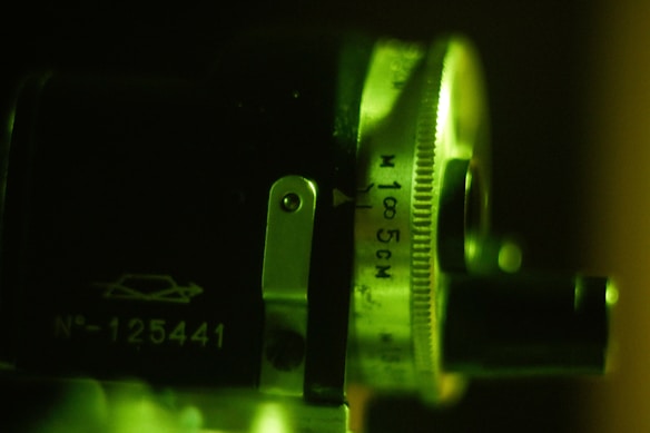 A close-up view of a mechanical or optical device featuring a metal dial with engraved measurements including '100 5CM'. Details like a small metal lever and a printed number 'N&deg;-125441' are visible. The image is illuminated with a greenish light, casting shadows and highlighting the textured surface.