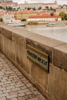 A stone walkway with a textured stone wall featuring a plaque labeled 'Karlův Most'. In the background, a river and boats are visible with an array of historic buildings displaying a mix of red and cream colors.