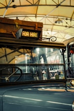 A vintage vehicle is inside a garage or station, with a suitcase on its roof. A sign reading 'FOR HIRE' is illuminated at the front. The background shows steel beams and a hanging light fixture, suggesting an industrial or rustic setting.