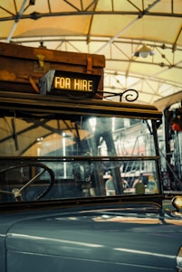 A vintage vehicle is inside a garage or station, with a suitcase on its roof. A sign reading 'FOR HIRE' is illuminated at the front. The background shows steel beams and a hanging light fixture, suggesting an industrial or rustic setting.