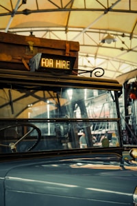 A vintage vehicle is inside a garage or station, with a suitcase on its roof. A sign reading 'FOR HIRE' is illuminated at the front. The background shows steel beams and a hanging light fixture, suggesting an industrial or rustic setting.