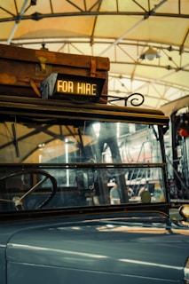 A vintage vehicle is inside a garage or station, with a suitcase on its roof. A sign reading 'FOR HIRE' is illuminated at the front. The background shows steel beams and a hanging light fixture, suggesting an industrial or rustic setting.