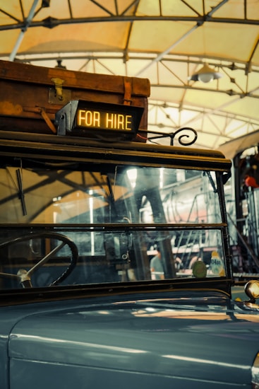 A vintage vehicle is inside a garage or station, with a suitcase on its roof. A sign reading 'FOR HIRE' is illuminated at the front. The background shows steel beams and a hanging light fixture, suggesting an industrial or rustic setting.