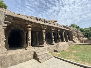A rock-cut temple carved into a cliff face, featuring intricately detailed stone pillars and sculptures of deities. The temple fa&ccedil;ade includes several columns with elaborate carvings, leading into a darkened interior. The background includes clear skies with scattered clouds and lush greenery surrounding the structure.