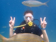 A scuba diver wearing a mask and breathing apparatus is underwater, holding up two peace signs. A fish swims above their head, and another fish is visible in the foreground. The background is a vibrant blue, indicating clear ocean water.