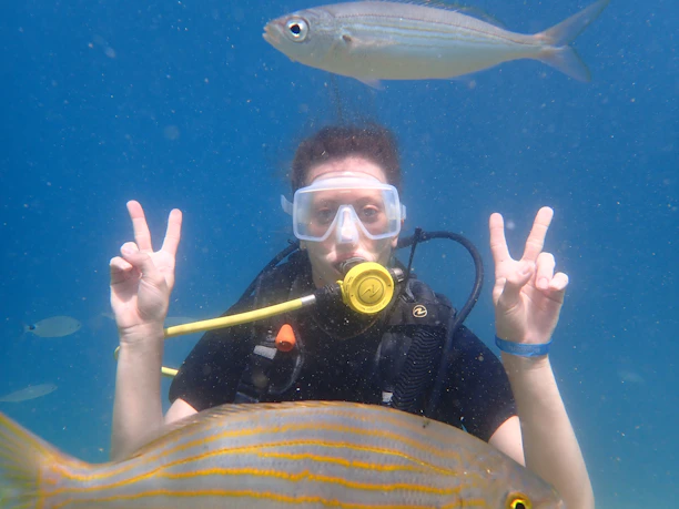 A smiling diver emerging from the ocean, holding up a peace sign with a bright blue sky behind.