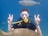 A scuba diver wearing a mask and breathing apparatus is underwater, holding up two peace signs. A fish swims above their head, and another fish is visible in the foreground. The background is a vibrant blue, indicating clear ocean water.