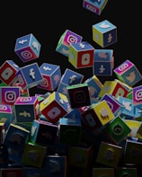 A collection of colorful 3D cubes displaying logos of various social media platforms, including Facebook, Instagram, Twitter, YouTube, and others. The cubes appear to be falling and tumbling over each other against a dark background.