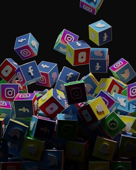 A collection of colorful 3D cubes displaying logos of various social media platforms, including Facebook, Instagram, Twitter, YouTube, and others. The cubes appear to be falling and tumbling over each other against a dark background.