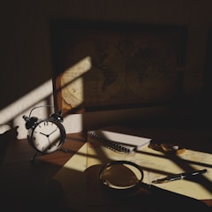 A dimly lit desk with an antique alarm clock showing seven o'clock, a spiral notebook, a magnifying glass, and a pen. A vintage world map on the wall casts a shadow over the workspace, creating a mysterious and contemplative atmosphere.