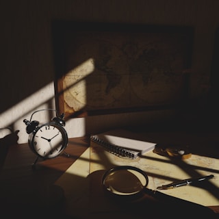 A dimly lit desk with an antique alarm clock showing seven o'clock, a spiral notebook, a magnifying glass, and a pen. A vintage world map on the wall casts a shadow over the workspace, creating a mysterious and contemplative atmosphere.